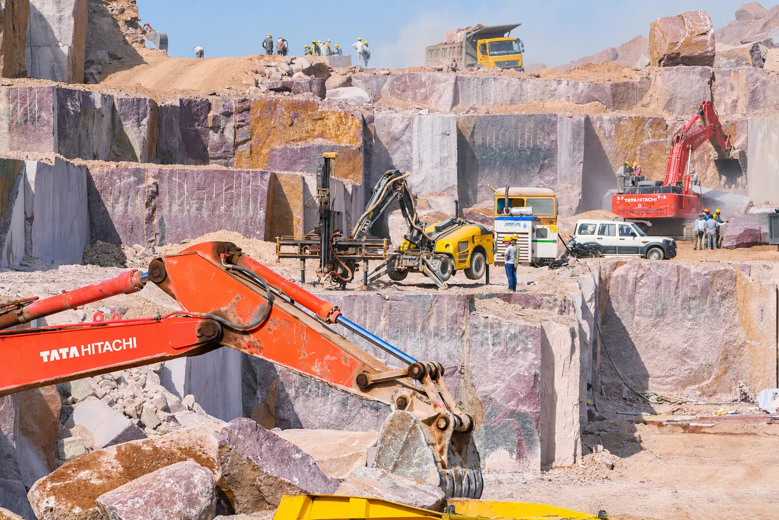 Working pink granite quarry with an excavator, a drill rig, workers and trucks on the benches